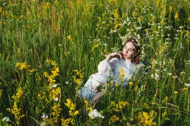 Beyaz elbiseli, başında çelenk olan güzel bir kadın çiçeklerle bir tarlada yatıyor. Güzellik, özgür yaşam ve doğallık kavramı.