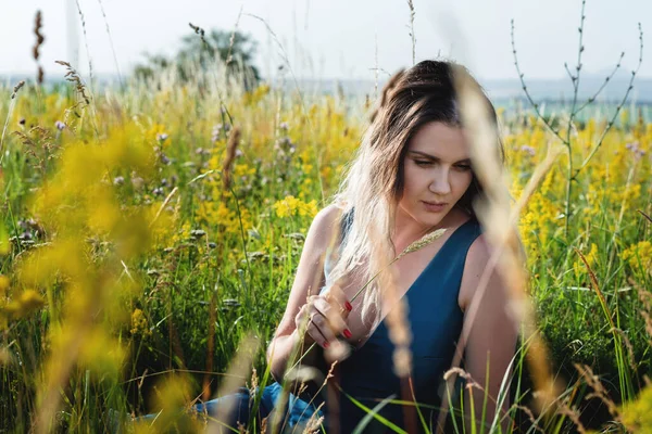 Uma mulher loira bonita em um vestido longo azul que se senta em um campo em flores. O conceito de beleza, vida livre e naturalidade — Fotografia de Stock