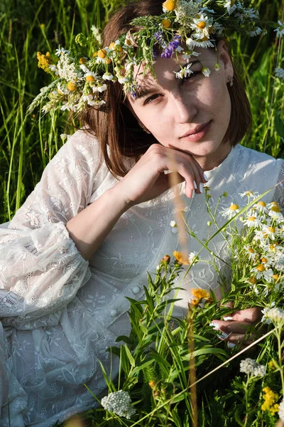 Krásná žena v bílých šatech ve stylu boho s věncem na hlavě leží na poli v květinách. Koncept krásy, volného života a přirozenosti — Stock fotografie