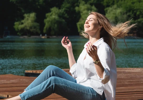 Uma mulher bonita e sexy, com cabelos longos em camisa branca e jeans, passa um tempo na margem do lago, cercada por florestas e montanhas. Conceito de descanso e relaxamento — Fotografia de Stock