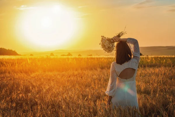 Schöne Frau in einem weißen Kleid mit einem Blumenstrauß in einem Weizenfeld bei Sonnenuntergang. Kostenloses Lifestylekonzept — Stockfoto