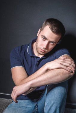 Mavi kotlu ve polo tişörtlü genç çekici bir adam stüdyonun duvarının yanında yerde oturuyor. Gençlik ve çekicilik kavramı