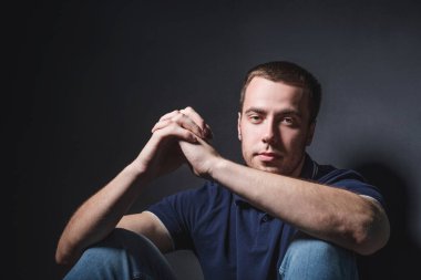 Mavi kotlu ve polo tişörtlü genç çekici bir adam stüdyonun duvarının yanında yerde oturuyor. Gençlik ve çekicilik kavramı