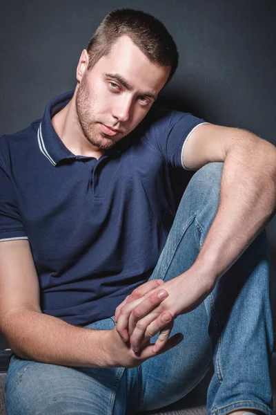 Joven hombre atractivo en jeans azules y una camisa de polo se sienta en el suelo cerca de la pared en el estudio. Concepto de juventud y atractivo — Foto de Stock