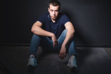 Mavi kotlu ve polo tişörtlü genç çekici bir adam stüdyonun duvarının yanında yerde oturuyor. Gençlik ve çekicilik kavramı