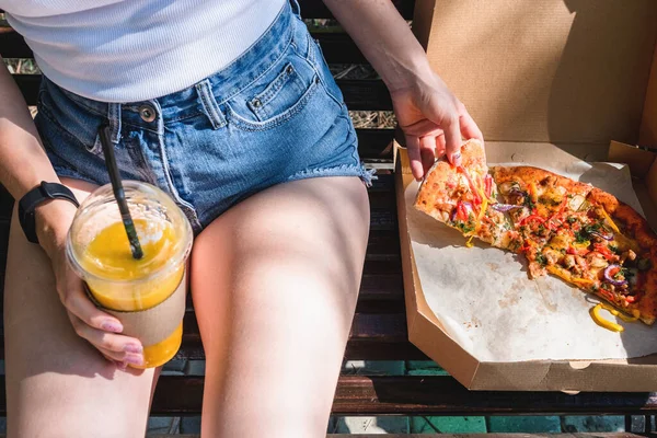 Güzel, genç bir kadın sokakta pizza ve patates kızartması yiyor. Fast food, yemek teslimatı ve doğada öğle yemeği kavramı.