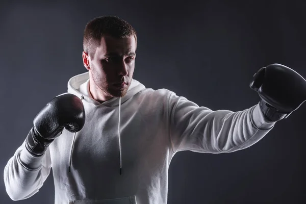 Jeune homme en gants de boxe et un sweat-shirt blanc sur fond sombre dans le studio. Le concept de victoires et de batailles — Photo