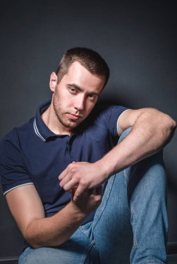 Mavi kotlu ve polo tişörtlü genç çekici bir adam stüdyonun duvarının yanında yerde oturuyor. Gençlik ve çekicilik kavramı