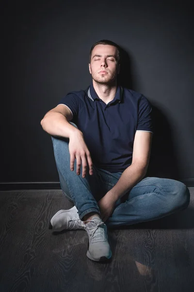 穿着蓝色牛仔裤和马球衫的年轻而有魅力的男人坐在工作室墙边的地板上。青年与魅力概念 — 图库照片