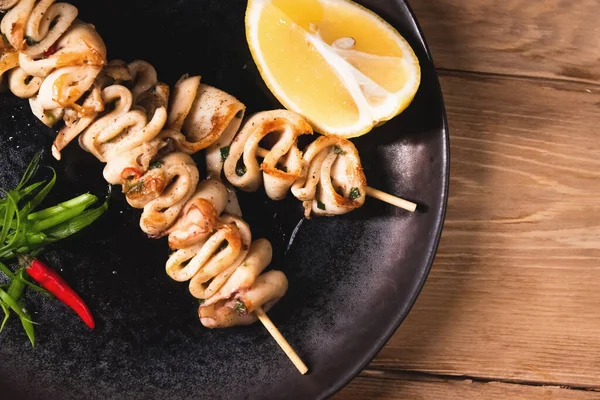 Fried squid bbq on skewers with lemon lies on a dark plate. Restaurant food and service concept — Stock Photo, Image