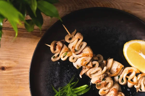 Fried squid bbq on skewers with lemon lies on a dark plate. Restaurant food and service concept — Stock Photo, Image