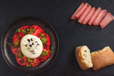 Burrata peyniri salatası, domates ve pesto sosu siyah bir tabakta et ve ekmeğin arasında. Çiftlik yemeği ve sofra kurma konsepti