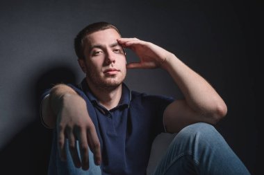 Mavi kotlu ve polo tişörtlü genç çekici bir adam stüdyonun duvarının yanında yerde oturuyor. Gençlik ve çekicilik kavramı