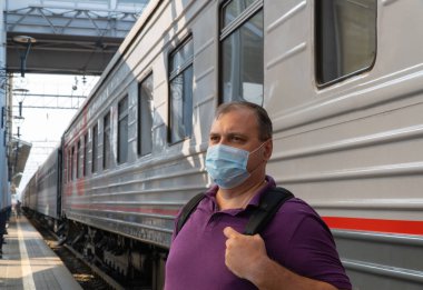 Tren vagonunun yanında maskeli bir adam var. Salgın sırasında seyahat kavramı. Koruyucu tıbbi maske takmanın kalıcı şekli. Cerrahi koruyucu maske. Tıbbi solunum bandajı yüzü.