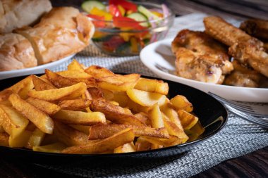 Siyah tabakta patates kızartması. Arka planda tavuk kanatları ve salata vardır.