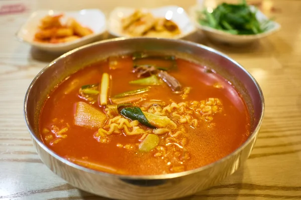 Kore kimchi ramen bir paslanmaz çelik kasede servis. Bulanık arka planda üç Kore gıda yan yemekler vardır