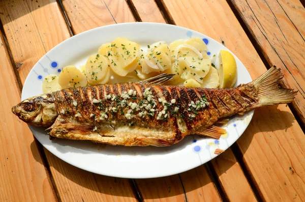 Grilled fresh fish in restaurant. Tasty and delicious fish with potatoes, lemon and garlic on plate on wooden table