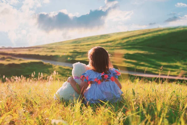 Uma menina em um vestido de 3 anos está sentada na grama com um ursinho na grama e abraçando. Conceito - amizade e amor à natureza. Uma criança em um prado com um brinquedo desfruta da paisagem . — Fotografia de Stock