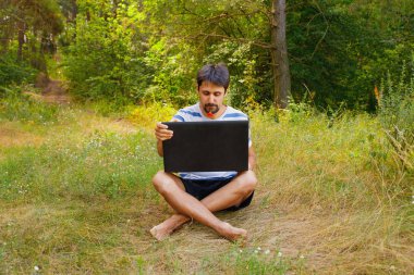 Genç bir adam ağzında bir çiçek ile çim oturur ve bir dizüstü bilgisayarda yazışmaçalışır.