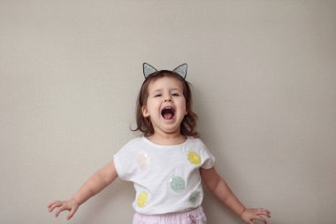 Beyaz tişörtlü, pembe şortlu neşeli küçük bir kızın yarım yamalak uzunluğu bej bir arka planda kolları açık çığlıklar atıyor.