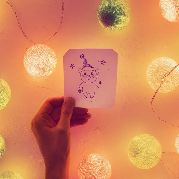Mano femenina sostiene un papel de cerdito dibujado sobre papel, hecho a mano, alrededor de la guirnalda luminosa de Año Nuevo, primer plano — Foto de Stock