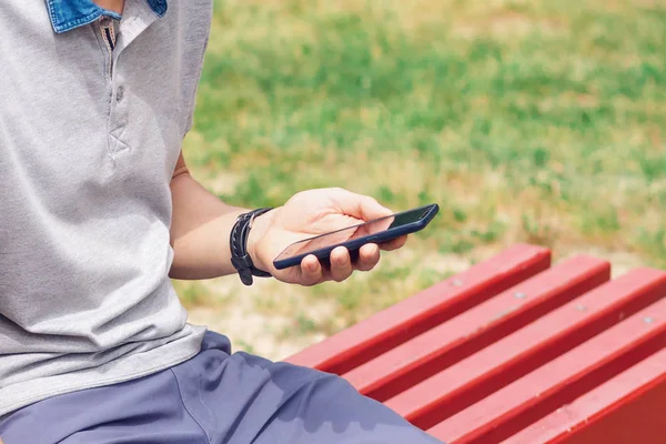 L'homme est assis sur un banc et utilise un smartphone avec un écran gris dans la rue de la ville, gros plan — Photo