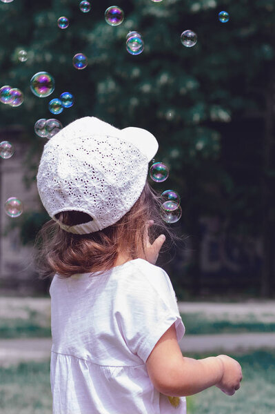 Белая девушка в белой футболке и кепке на фоне травы играет с пузырьками воздуха. Вид сзади
