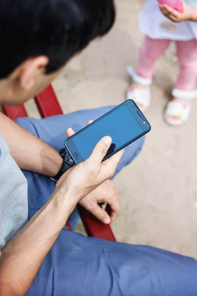 Un homme s'assoit sur le terrain de jeu et utilise un smartphone, ne fait pas attention à sa fille — Photo