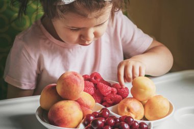 3 yıllık beyaz tişörtlü küçük kız masada taze meyve yiyor..