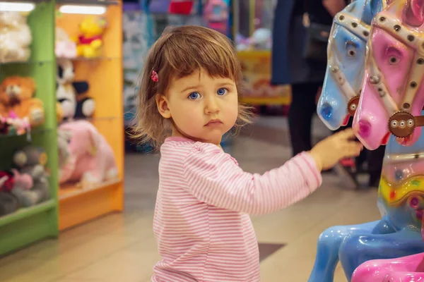 little girl of Caucasian appearance in jeans and T-shirt with surprised expression stroking toy horse in fun center