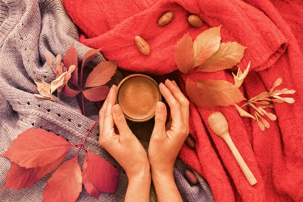 Frauenhände in der Hand eine Tasse veganen Kakaos, herbstlicher Hintergrund mit rotgrauem Karo und gelben Blättern, Tageslicht, Flatlay, Draufsicht. Warmgetränkekonzept. — Stockfoto