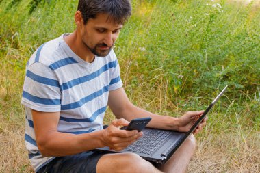 Sakallı genç bir adam çimlerin üzerinde oturur ve bilgisayarın başında çalışır. Kavram - serbest çalışmak zevktir.