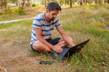 Sakallı genç bir adam çimlerin üzerinde oturur ve bilgisayarın başında çalışır. Kavram - serbest çalışmak zevktir.