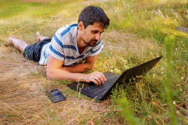 Sakallı genç bir adam çimlerin üzerinde oturur ve bilgisayarın başında çalışır. Kavram - serbest çalışmak zevktir.