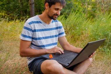 Sakallı genç bir adam çimlerin üzerinde oturur ve bilgisayarın başında çalışır. Kavram - serbest çalışmak zevktir.
