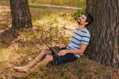 Sakallı genç bir adam çimlerin üzerinde oturur ve bilgisayarın başında çalışır. Kavram - serbest çalışmak zevktir.