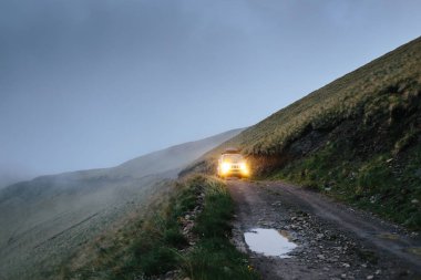 Off-road car on road trip in mountains. Compact SUV goes uphill on narrow dangerous stony road in the fog with headlights on clipart