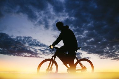 Bisikletçi dramatik bir bulutlu gökyüzü karşı baş lambalı karanlıkta açık havada dağ bisikleti binme. Düşük açılı görünümü ile hareket bulanık siluet. Bisiklet aydınlatma donanımları. Aktif yaşam tarzı kavramı