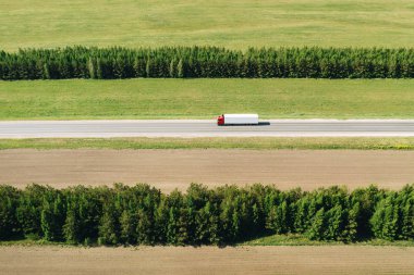 Güneşli bir günde ağaçların iki satır arasında bir kırsal karayolu üzerinde beyaz kargo römork ile kırmızı yarı kamyon havadan çekim. Bir yaz sezonunda ulaşım