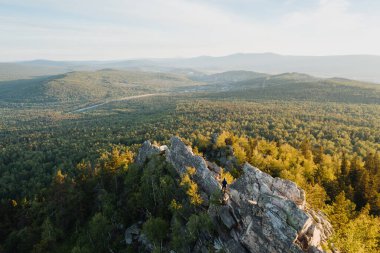 Kayaların tepesindeki adam, gün batımında yeşil ormanın arasında güzel vadiye tepeden bakan. Açık hava ve dağ yürüyüşü geçmişi. Chelyabinsk Oblastı 'ndaki Zlatoust yakınlarındaki doğa manzarası, Ural, Rusya