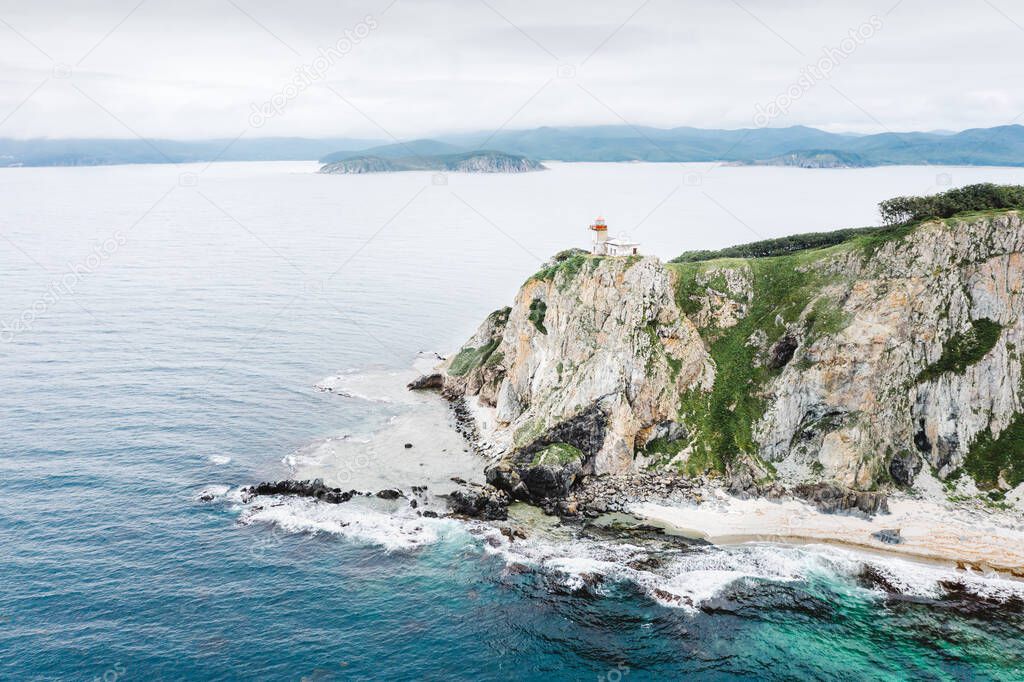 Lighthouse at green rocky coast of Cape Balyuzek, aerial drone view. Sea of Japan at Balyuzek Peninsula coastline in summertime. Seaside nature landscape in Primorsky Krai, Far East, Russia