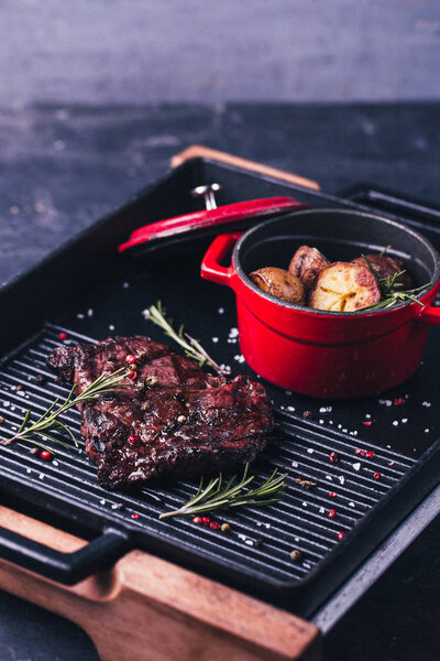  steak with rosemary and baked potatoes in red pan on black background