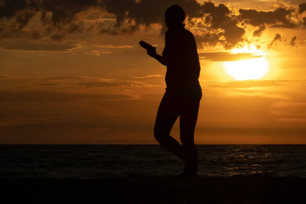 Contour image of a young woman against the background of the setting sun over the sea. A young woman walks along the beach, holding a phone in her hand. Blurred into motion.