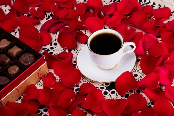 Closeup of cup of coffee and box of chocolates surrounded by red rose petals