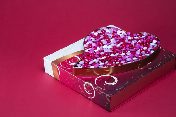 Heart shaped box filled with pink and white coated candy on top of a red white and gold gift box