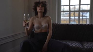 Elinde bir bardak beyaz şarap ile kanepede siyah kıvırcık saçlı bej sutyen ve mavi geniş pantolon giyen güzel bir kadın oturur, o gülümsüyor ve cam kamera hits
