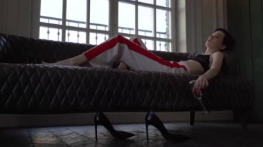 Kırmızı çizgiler ve siyah sütyen beyaz pantolon giymiş kısa mor saçlı bir kadın düşünceli karanlık bir kanepede yatıyor ve bir kadeh şampanya elinde tutan