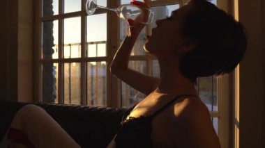 Kısa saç kesimi ve siyah saçlı, siyah sütyen giymiş bir kadınla siyah bir divan büyük pencerede bir kadeh şampanya, holding onun içme, şampanya vücuttaki damlayan ellerinde