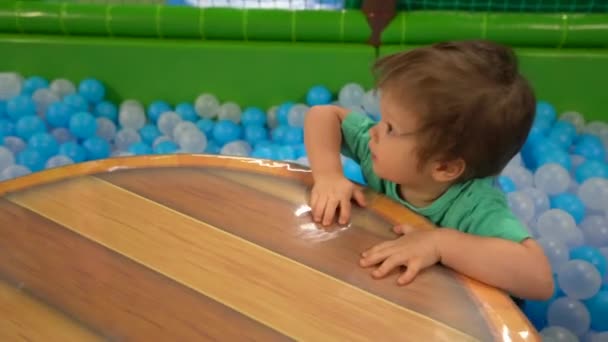 一个两三岁的男孩穿着绿松石 T恤和深蓝色裤子 一个人站在一个干池里 有白色和蓝色的球 然后妈妈帮忙去游泳池 — 图库视频影像