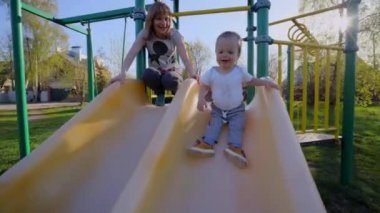 Bir anne ile küçük bir çocuk/çocuk parkta bir güneşli yaz gününde bebek sarı tepeden aşağı taşır yürür ve annesi izliyor